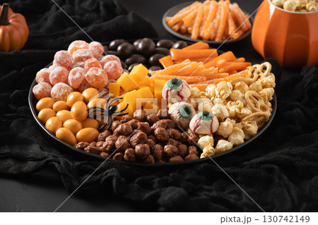 Halloween sweet charcuterie board with pumpkin, popcorn, orange chocolate candy, spider, crackers on black background. Scary decoration. Festive food for kids party. Close up. Halloween sweet charcuterie board with pumpkin, popcorn, orange chocolate candy, spider, crackers on black background. Scary decoration. Festive food for kids party. Close up. 130742149