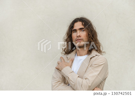 Handsome caucasian man, brunette with curly long hair wearing beige casual attire stands against neutral background looking at camera. Contemplation and introspection. Space for design. Handsome caucasian man, brunette with curly long hair wearing beige casual attire stands against neutral background looking at camera. Contemplation and introspection. Space for design. 130742501