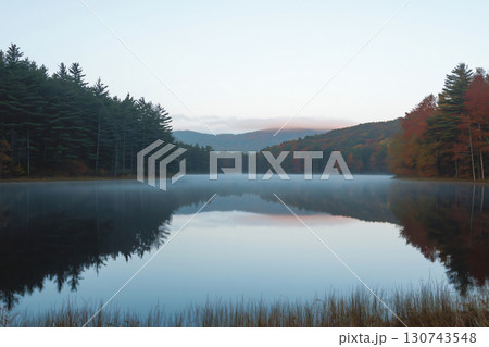 Tranquil lake reflecting the surrounding forest and sky with a thin layer of fog hovering above the water 130743548