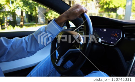 Male hand of businessman holds steering wheel while driving electrical vehicle at city. Businessperson operating electric car on urban road. Young man commuting to work at sunny day. Close up Male hand of businessman holds steering wheel while driving electrical vehicle at city. Businessperson operating electric car on urban road. Young man commuting to work at sunny day. Close up 130745331