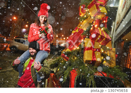a young woman sits on the shoulders of a bearded man and smiles against the backdrop of a red retro car with Christmas gifts on the roof and against the backdrop of snow a young woman sits on the shoulders of a bearded man and smiles against the backdrop of a red retro car with Christmas gifts on the roof and against the backdrop of snow 130745658
