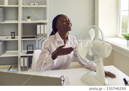 Young african american woman working at office using electric fan during heatwave in office. Young african american woman working at office using electric fan during heatwave in office. 130746289