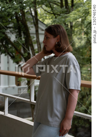 Side portrait of a woman standing on a balcony, touching her hair while looking away, surrounded by lush greenery and white classical architecture 130746540