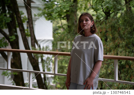 A thoughtful woman in a grey oversized T-shirt stands on a rainy balcony surrounded by green foliage and white architecture 130746541