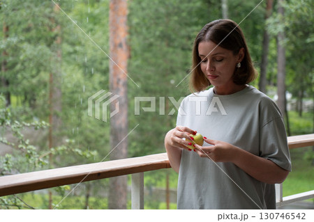 Young woman with short hair and red nails gently holds a green apple while standing on a porch during a summer rain in the forest 130746542