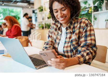 A focused young woman is sitting in a vibrant cafe, using both her smartphone and laptop 130747164