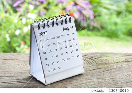 August 2027 spiral desk calendar on wooden surface with blurred outdoor greenery background. August 2027 spiral desk calendar on wooden surface with blurred outdoor greenery background. 130748010