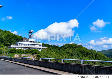 青空を背景に建つ布目ダム管理事務所とダム堰堤 130748874