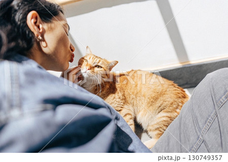 Affectionate Woman Leaning to Kiss Ginger Cat in Sunlight. 130749357