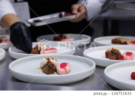plated desserts in professional restaurant kitchen. chef arranges chocolate cake, mousse, and artistic garnishes on plates. fine dining presentation, culinary art and gourmet food preparation. plated desserts in professional restaurant kitchen. chef arranges chocolate cake, mousse, and artistic garnishes on plates. fine dining presentation, culinary art and gourmet food preparation. 130751635