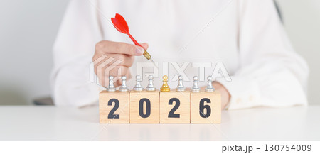 Hand holding red dart over people chess and 2026 year blocks. Goal, plan, action, target, strategy, leadership, New Year and marketing concept 130754009