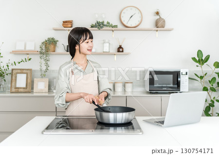 キッチンでパソコンを見ながら料理するエプロン姿の若いアジア人女性 キッチンでパソコンを見ながら料理するエプロン姿の若いアジア人女性 130754171