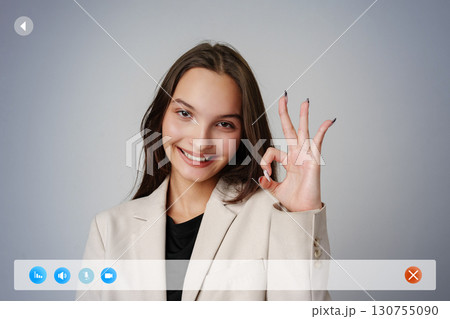 Young woman in a light suit making an okay gesture in a professional setting against a neutral background 130755090