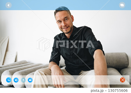 Young man smiling while sitting on a light gray couch in a modern living room during daylight hours Young man smiling while sitting on a light gray couch in a modern living room during daylight hours 130755606
