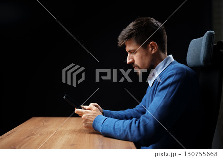 Man using smartphone while sitting at a wooden desk in a dimly lit room during evening hours 130755668