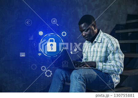 Man using laptop on stairs with digital security icons in a modern workspace during evening hours 130755881