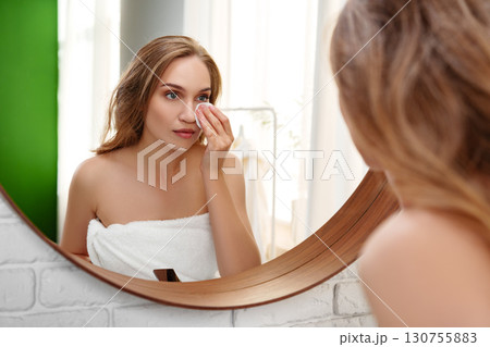 Woman applying skincare in front of round mirror in a bright bathroom after showering and wrapping her hair in a towel Woman applying skincare in front of round mirror in a bright bathroom after showering and wrapping her hair in a towel 130755883