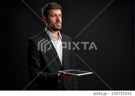Professional man in formal attire holds tablet while looking thoughtfully against dark background 130755932