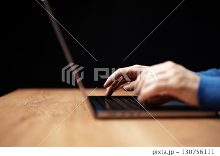 Person typing on a laptop at a wooden desk in a dimly lit environment during evening hours Person typing on a laptop at a wooden desk in a dimly lit environment during evening hours 130756111