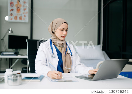 Beautiful physician doctor sit on table in office hospital. Attractive therapist female practitioner pharmacist smile and use tablet and laptop. 130756242
