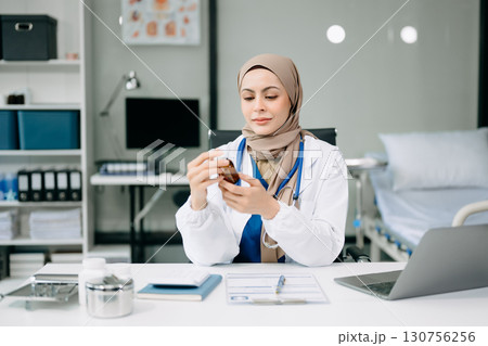 Beautiful physician doctor sit on table in office hospital. Attractive therapist female practitioner pharmacist smile and use tablet and laptop. 130756256