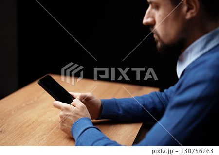 Man focused on smartphone at wooden table in dimly lit indoor space during evening hours 130756263