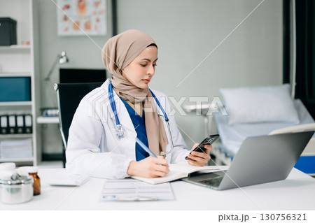 Beautiful physician doctor sit on table in office hospital. Attractive therapist female practitioner pharmacist smile and use tablet and laptop. 130756321