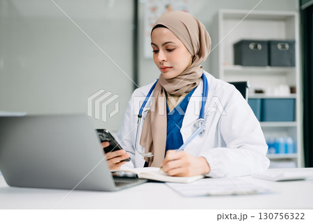 Beautiful physician doctor sit on table in office hospital. Attractive therapist female practitioner pharmacist smile and use tablet and laptop. Beautiful physician doctor sit on table in office hospital. Attractive therapist female practitioner pharmacist smile and use tablet and laptop. 130756322