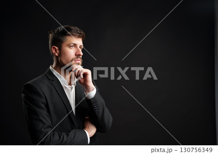 Businessman in formal suit pondering ideas against dark background in a professional setting Businessman in formal suit pondering ideas against dark background in a professional setting 130756349