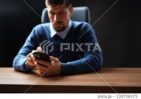 Man in sweater focused on smartphone at desk in dimly lit office environment during evening hours Man in sweater focused on smartphone at desk in dimly lit office environment during evening hours 130756375