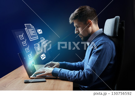 Professional man working on laptop with virtual documents and messages in a modern office setting at night 130756592