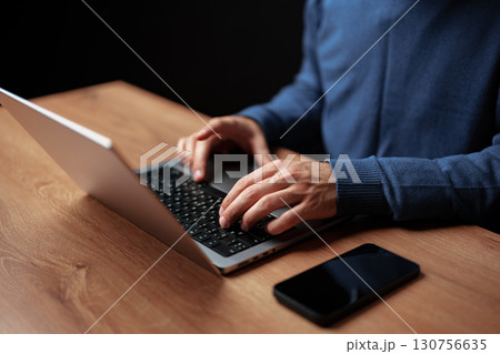 Typing on laptop at wooden desk during evening hours with smartphone nearby 130756635