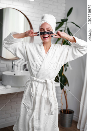 Woman enjoying a spa day at home while smiling and posing with facial mask in a modern bathroom Woman enjoying a spa day at home while smiling and posing with facial mask in a modern bathroom 130756676