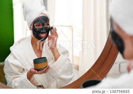 Woman applying face mask in bathrobe while smiling in front of mirror in bright room 130756685