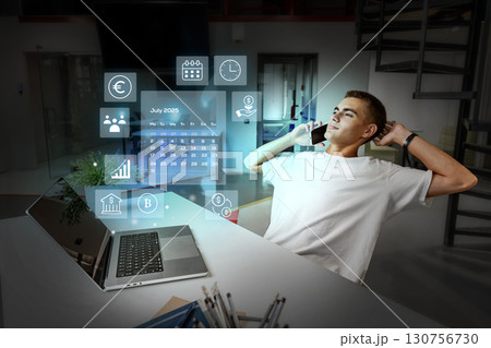 Young man engages in remote work while using a laptop and phone in a futuristic office setting during the evening hours Young man engages in remote work while using a laptop and phone in a futuristic office setting during the evening hours 130756730
