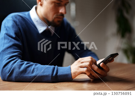 Man using smartphone while sitting at a wooden desk in a modern office during daytime Man using smartphone while sitting at a wooden desk in a modern office during daytime 130756891