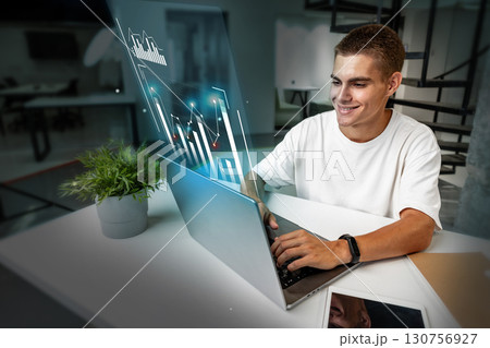 Young man working on laptop with digital graphs in modern workspace during daytime 130756927