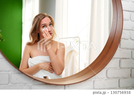 Woman applying skincare in a bright bathroom while looking in a round mirror 130756938