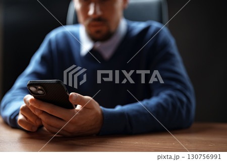 Man using smartphone while sitting at a wooden desk during a work session in an office environment Man using smartphone while sitting at a wooden desk during a work session in an office environment 130756991