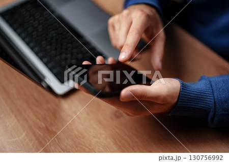 Person using smartphone while working at a wooden desk with a laptop nearby during the day Person using smartphone while working at a wooden desk with a laptop nearby during the day 130756992