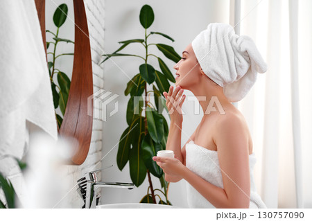 Woman applying facial cream in a serene bathroom setting with natural light during the day 130757090