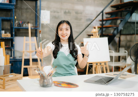 Cheerful female artist painting in a vibrant studio with brushes, a palette, and a blank canvas. 130758275