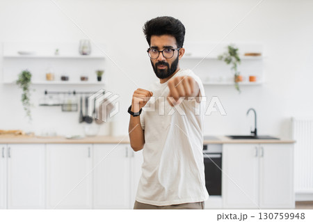 Young adult man with black hair and glasses performing boxing moves in home setting. Casual outfit in a bright kitchen emphasizes focus, determination, and fitness in a cozy and familiar indoor space. Young adult man with black hair and glasses performing boxing moves in home setting. Casual outfit in a bright kitchen emphasizes focus, determination, and fitness in a cozy and familiar indoor space. 130759948