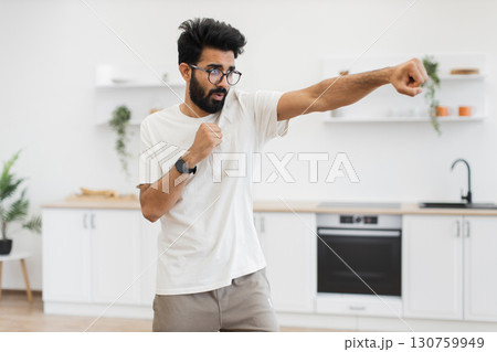 Young adult man performing boxing practice alone in a bright home kitchen environment. Wearing casual outfit, displaying enthusiasm and focus, creating dynamic and energetic scene in domestic setting. Young adult man performing boxing practice alone in a bright home kitchen environment. Wearing casual outfit, displaying enthusiasm and focus, creating dynamic and energetic scene in domestic setting. 130759949