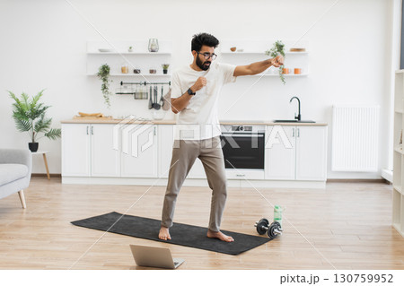 Middle-aged man doing fitness boxing exercises in bright kitchen, barefoot, using yoga mat. Middle-aged man doing fitness boxing exercises in bright kitchen, barefoot, using yoga mat. 130759952