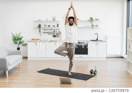 Adult male practicing yoga in living room, demonstrating fitness and health in modern home setting. Adult male practicing yoga in living room, demonstrating fitness and health in modern home setting. 130759979