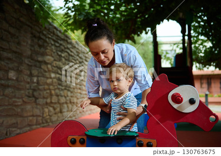 Mother Helping Son on Playground Rocking Horse in Outdoor Play Area Mother Helping Son on Playground Rocking Horse in Outdoor Play Area 130763675