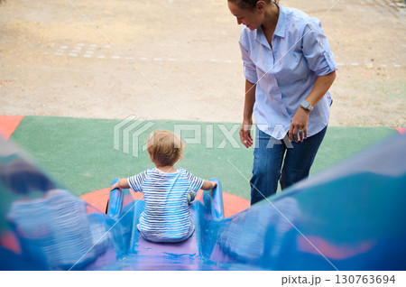 Mother Guiding Her Toddler on a Slide at a Playground 130763694