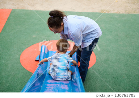 Mother Assisting Toddler on Playground Slide for Fun and Safety Mother Assisting Toddler on Playground Slide for Fun and Safety 130763705