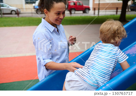 Mother Assisting Young Child Climbing Playground Slide on a Sunny Day Mother Assisting Young Child Climbing Playground Slide on a Sunny Day 130763717